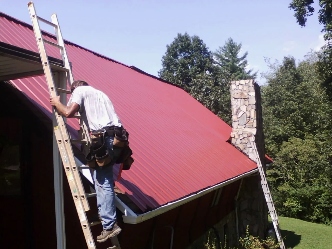 Roofer on a ladder beside a red metal roof and stone chimney — Soffit & Fascia Repair by Roofing Brothers in Mellott